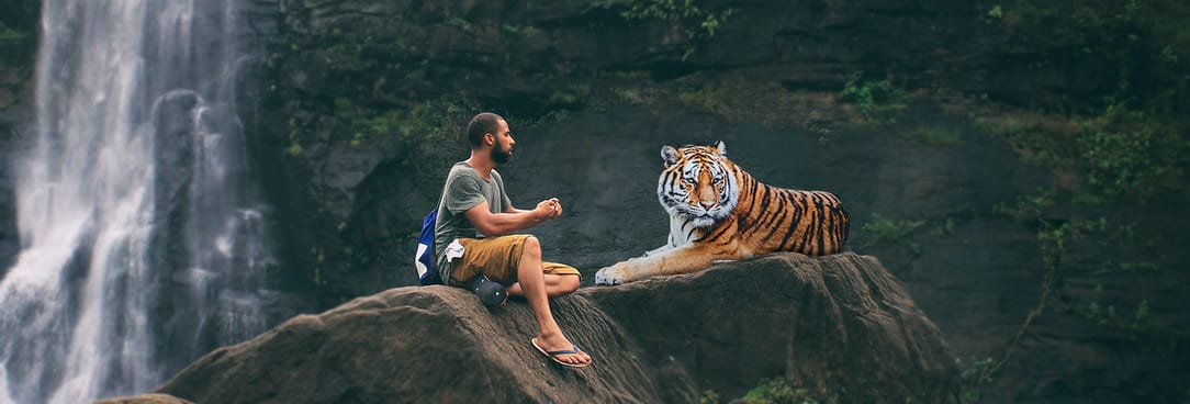 A man sitting on a large rock, with a tiger laying next to him. The man seems to be talking to the tiger. The tiger has its face averted.